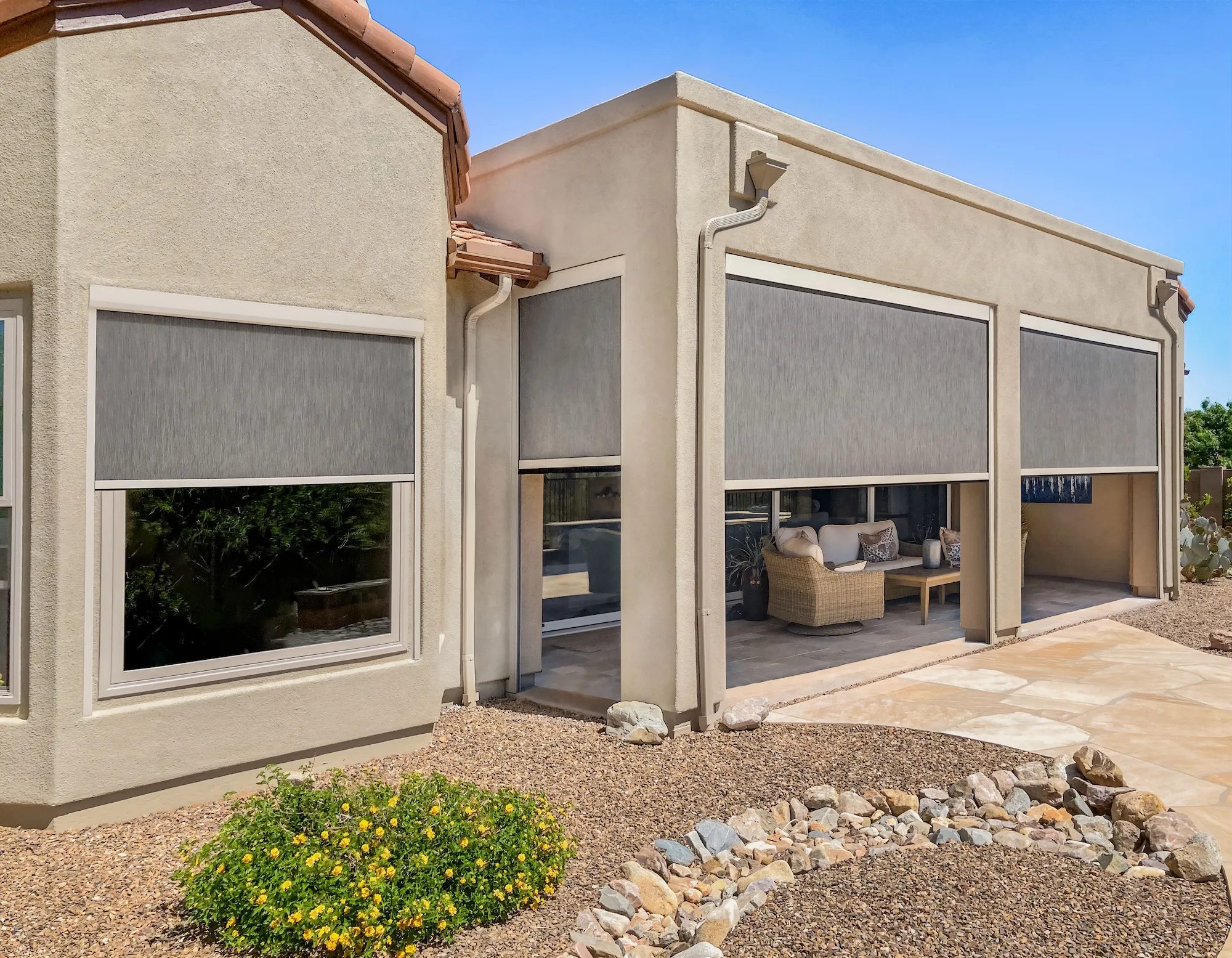 Modern beige stucco house with multiple windows and patio openings covered by gray fabric outdoor shades, with a gravel yard and a small flowering bush.