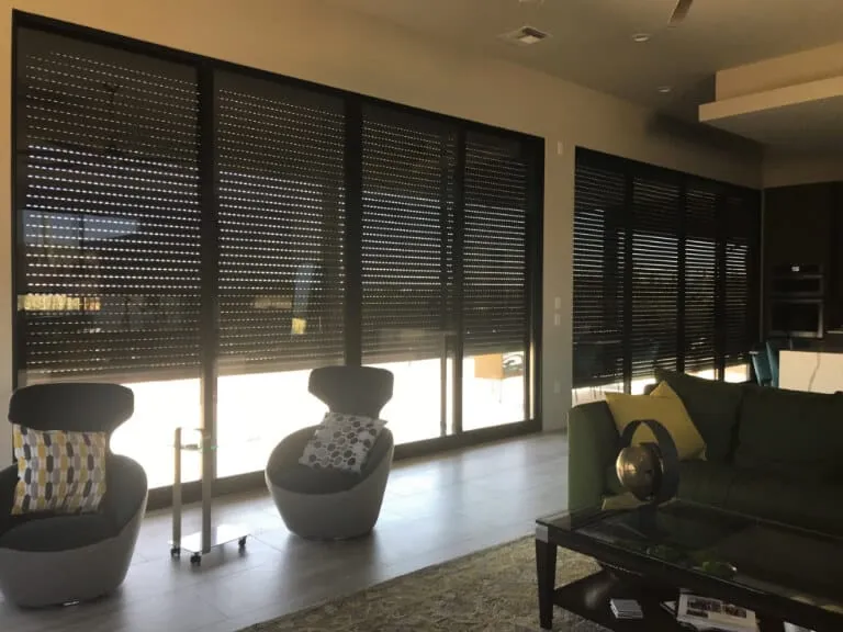 Modern living room with large windows and dark blinds, featuring two stylish chairs and a green sofa.