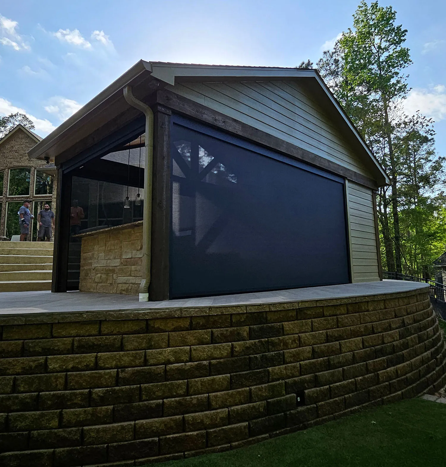 Outdoor covered patio with a dark roll-down screen, stone retaining wall, and two men talking near stairs in the background.