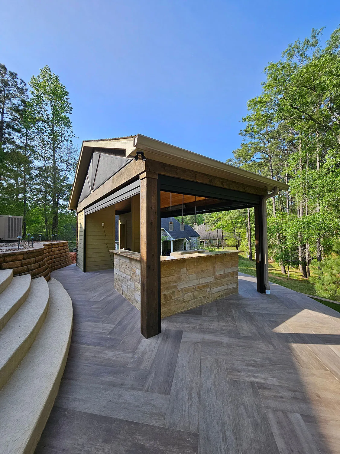 Outdoor covered patio bar area with stone counter, wooden beams, and gray patterned flooring, surrounded by trees under clear blue sky.