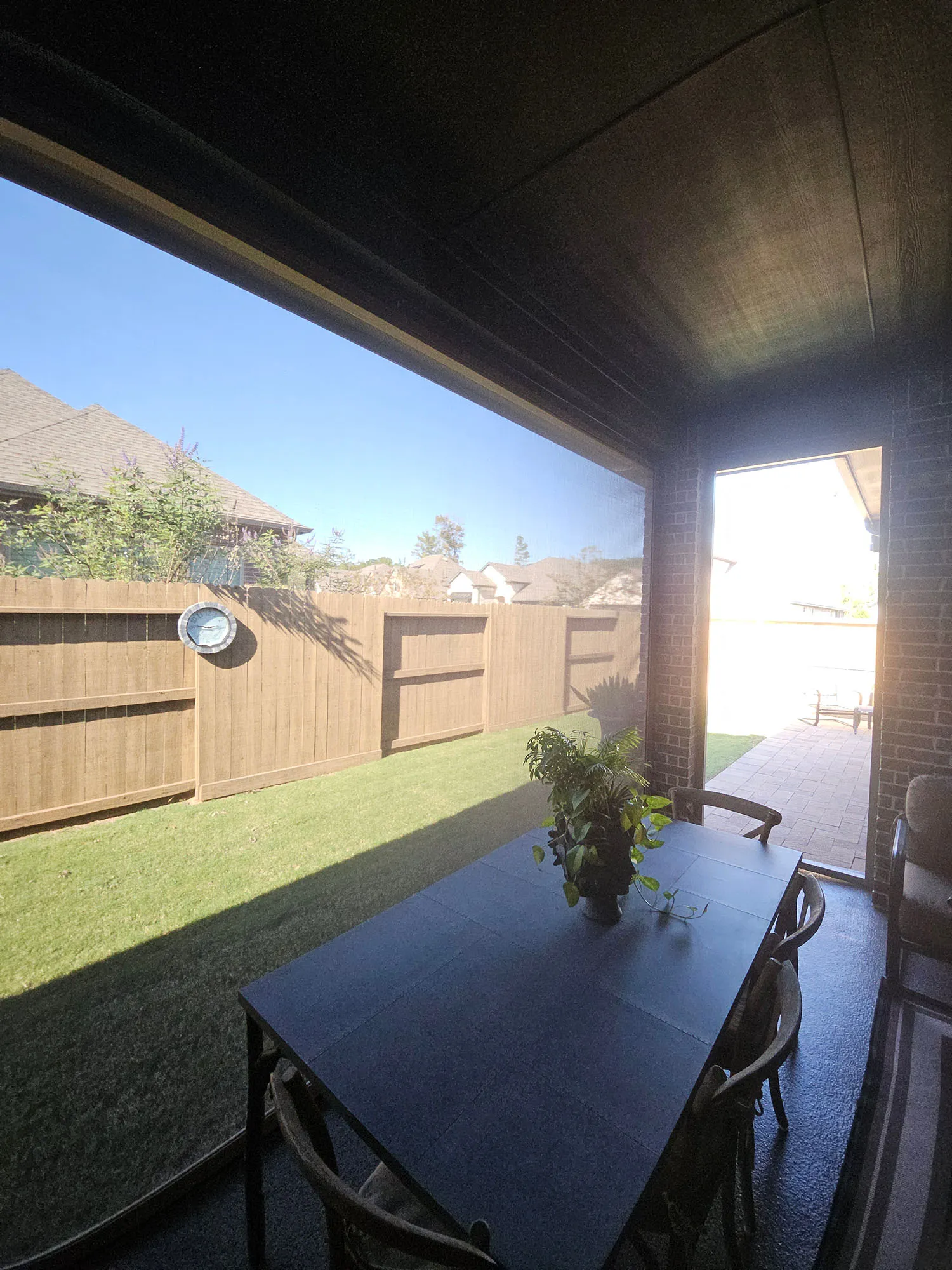 Covered patio with a black table, four chairs, and a potted plant, overlooking a green lawn and wooden fence under a clear blue sky.