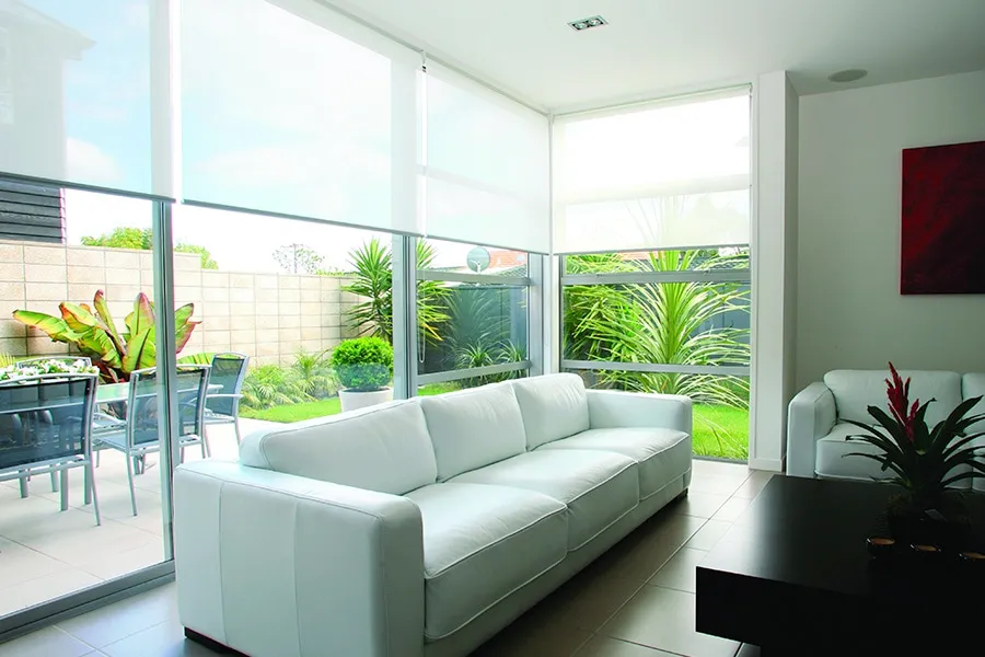 Bright modern living room with white leather sofas, large corner windows with roller blinds, and a view of a garden patio with outdoor seating.
