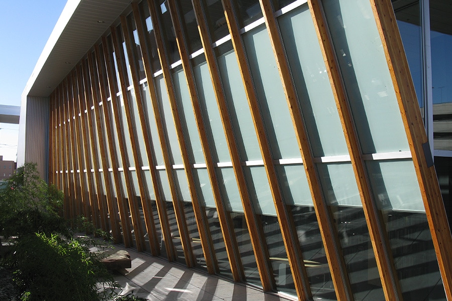 Modern building facade with large angled glass windows supported by vertical wooden beams casting shadows on the walkway below.