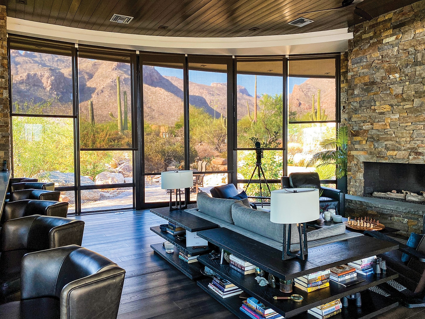 Modern living room with large windows showing desert mountains, leather chairs, a stone fireplace, and a chessboard on a wooden coffee table.