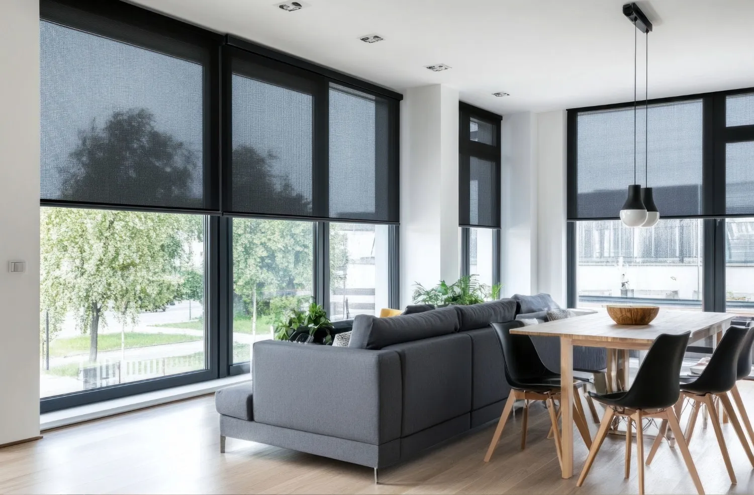 Modern living and dining area with large windows covered by black roller blinds, a grey sectional sofa, wooden dining table, and black chairs.