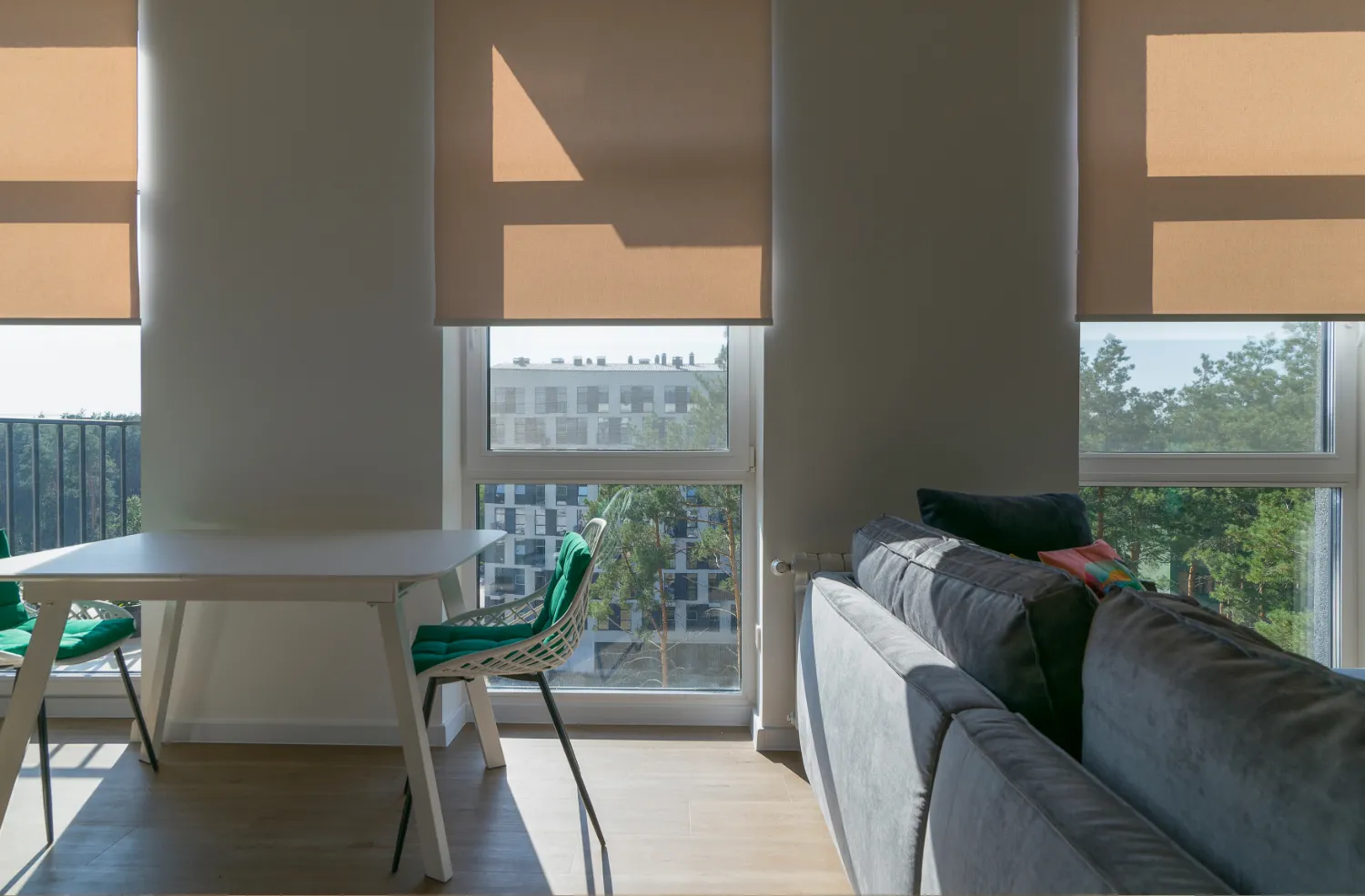 Modern living space with a white table and green cushioned chairs beside large windows with beige blinds and a gray sofa.