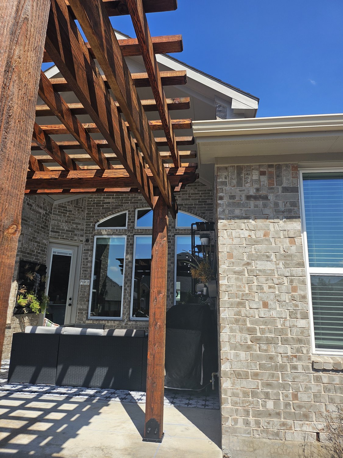 Wooden pergola casting shadows on a patio with brick walls, outdoor sofa, and large windows under a clear blue sky.