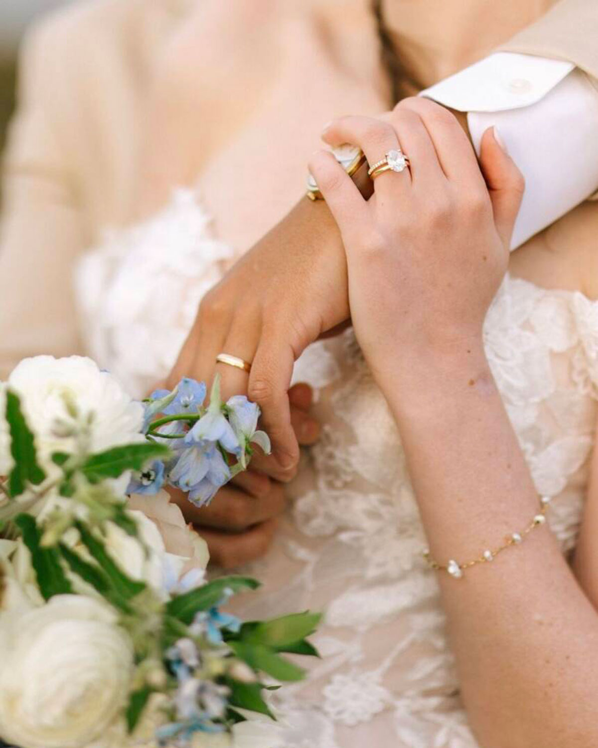 Two hands with wedding rings gently holding each other over a white lace dress and blue flowers.