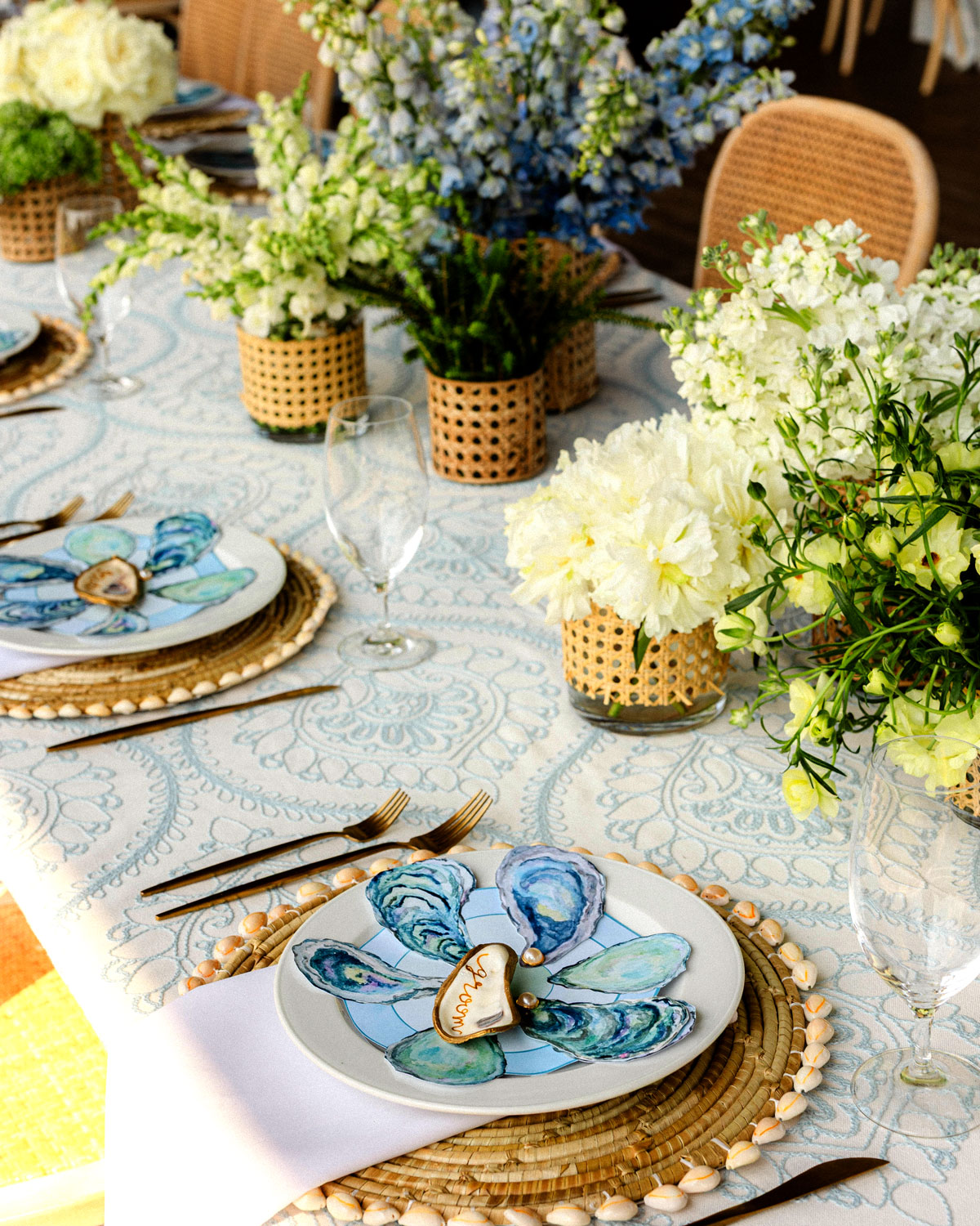Table setting with rattan chargers, oyster shell place card, blue floral plates, and white flowers.