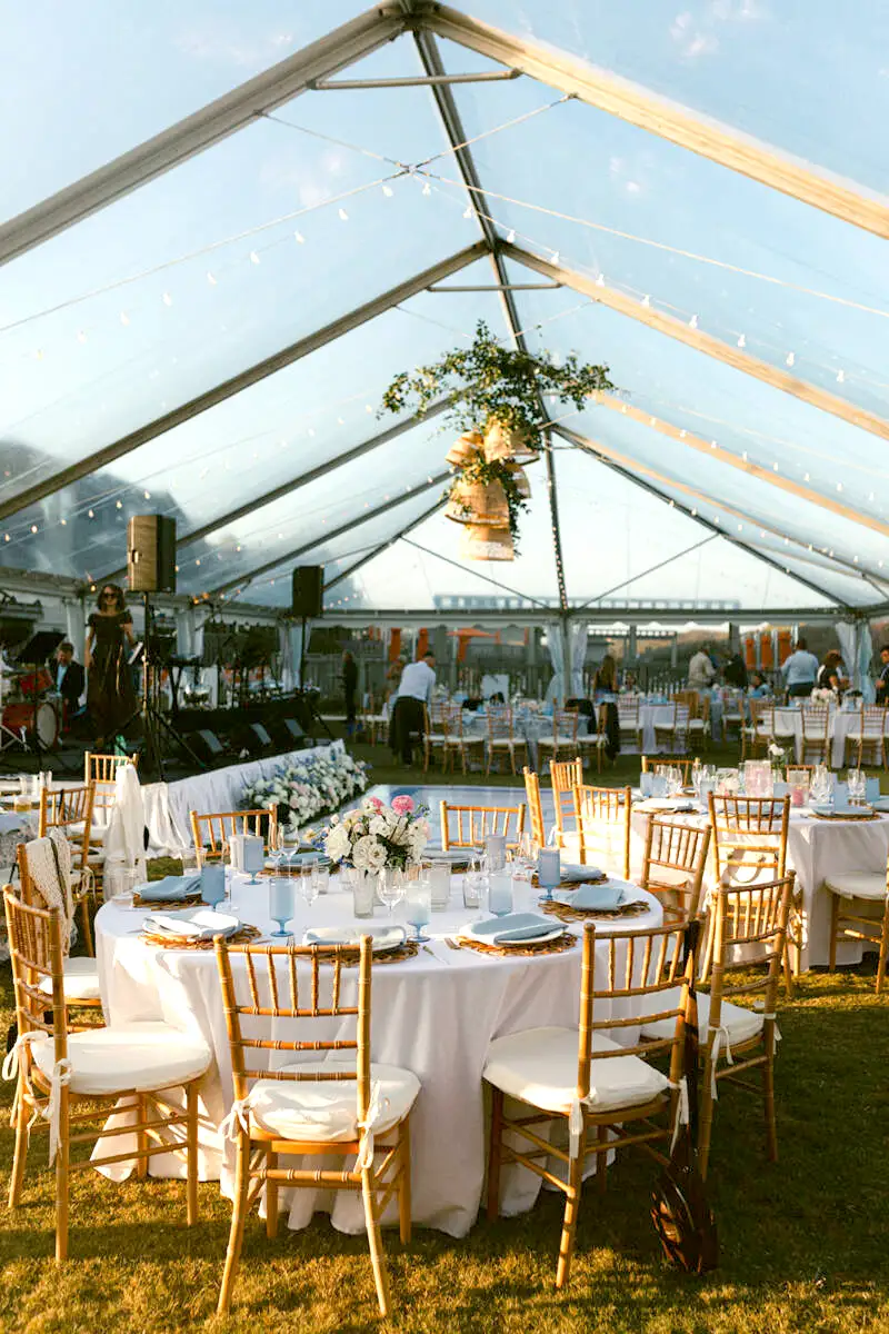 Elegant outdoor event setup under a transparent tent with round tables and wooden chairs on grass.