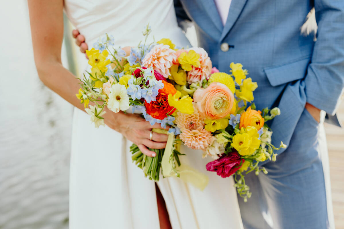 Bride and groom holding a vibrant bouquet of mixed yellow, peach, red, and blue flowers.