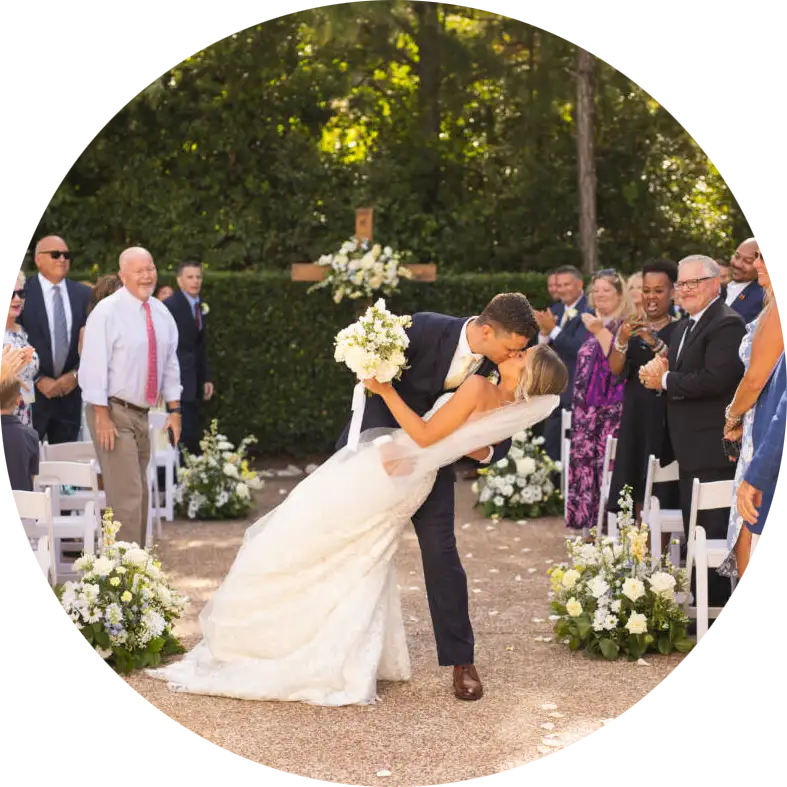 Bride and groom sharing a kiss as guests applaud at an outdoor wedding ceremony aisle decorated with white flowers.