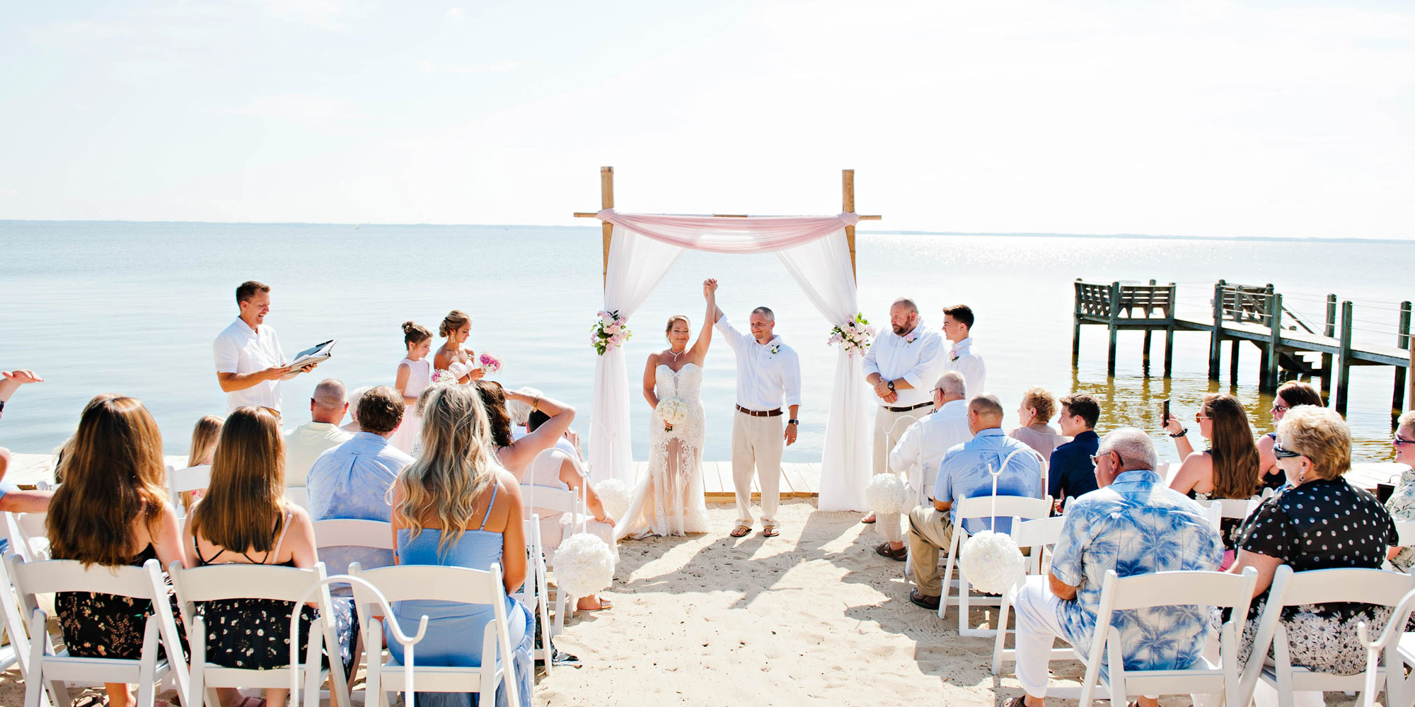 Bride and groom holding hands under decorated arch on beach with seated guests watching.