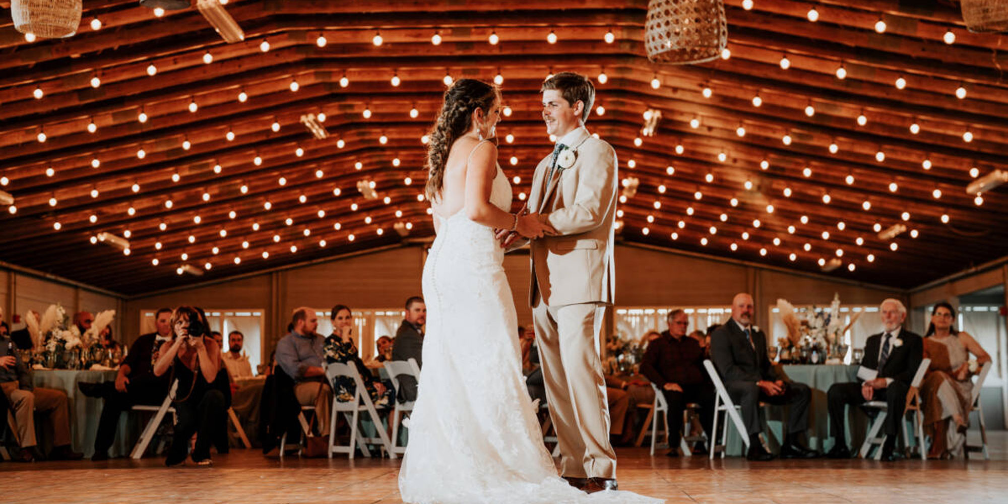 Bride and groom holding hands and dancing under string lights with seated guests watching.