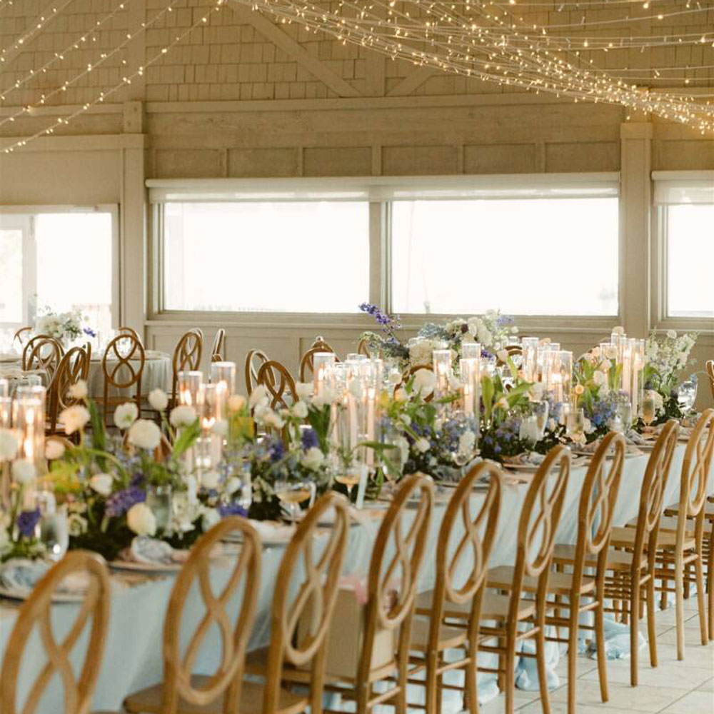 Long banquet table with wooden chairs, candles, and floral arrangements under string lights.