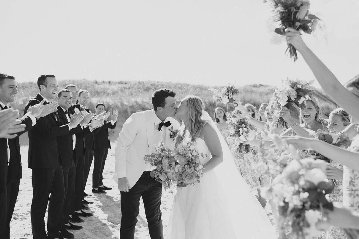 Bride and groom kissing on a beach while bridal party in dresses and tuxedos clap and cheer.