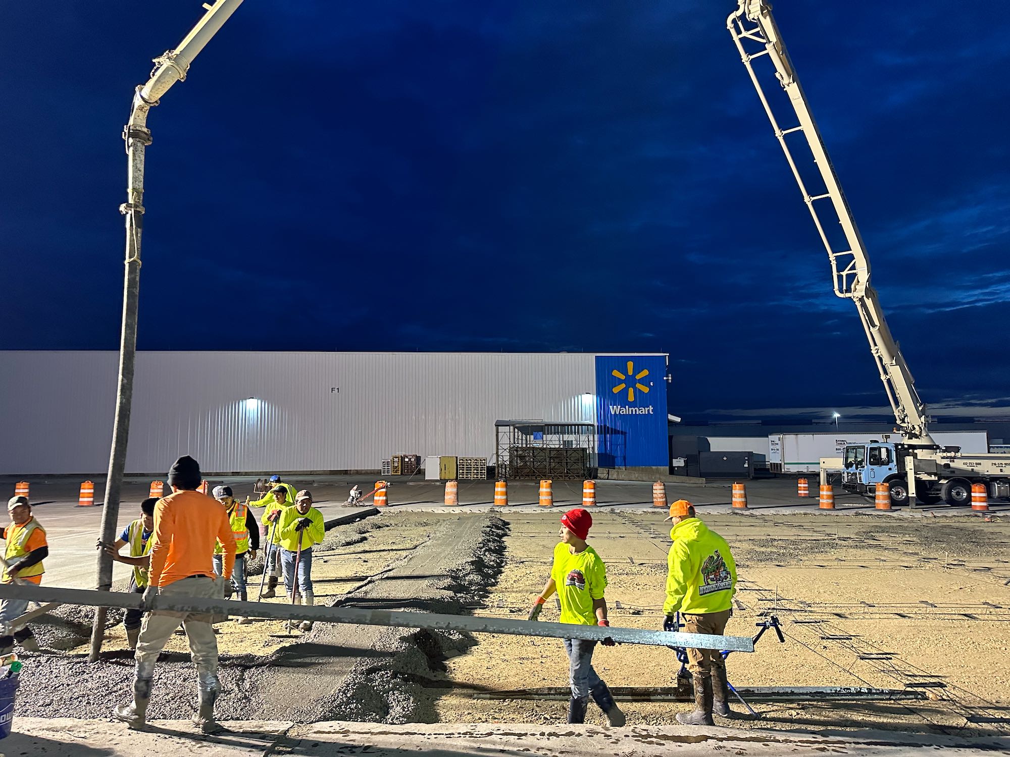 Construction workers pouring and leveling concrete at a Walmart site during early evening with dark blue sky.