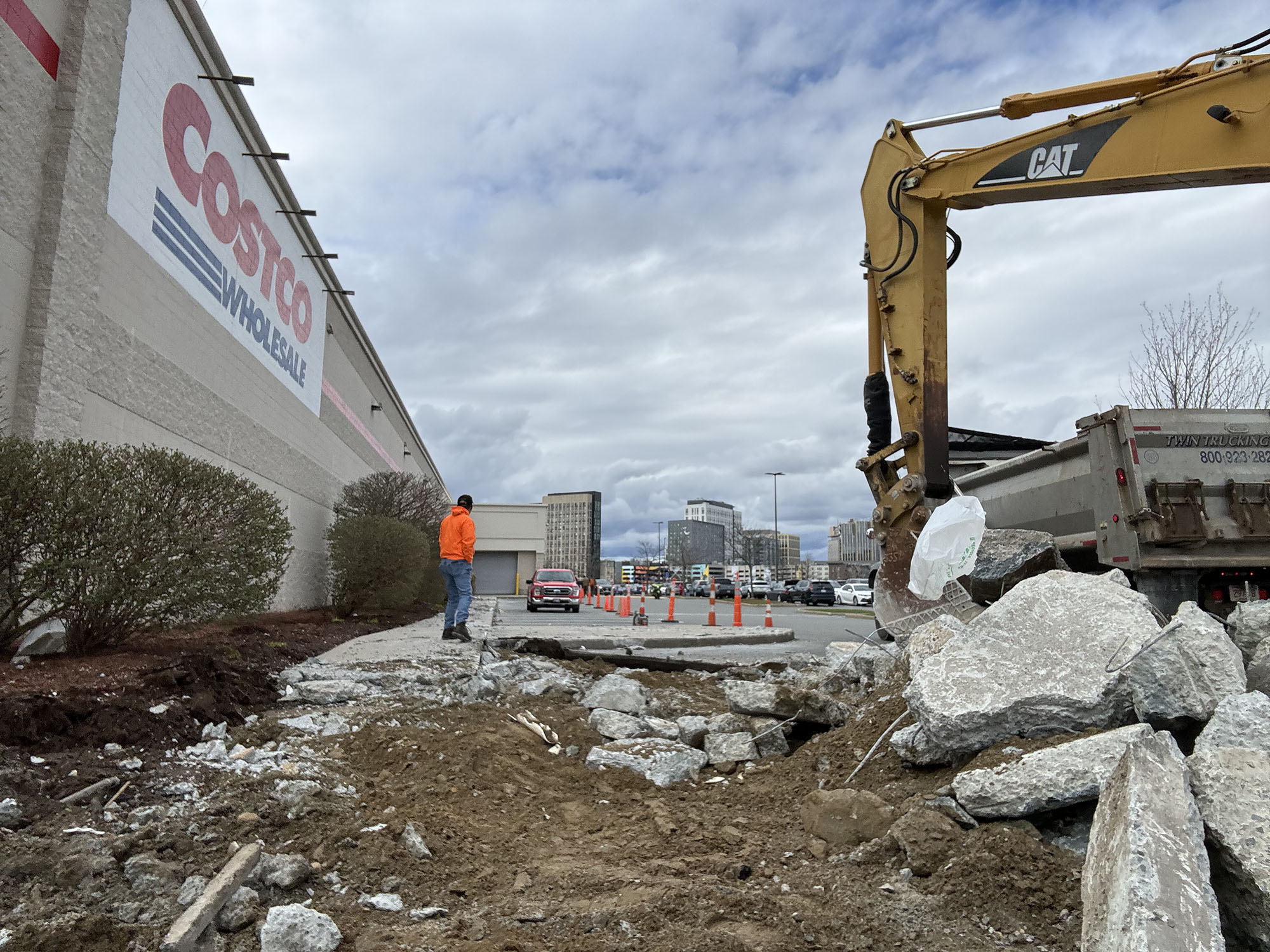 Construction work with an excavator digging up concrete near a Costco Wholesale store, with a worker in an orange jacket standing nearby and a parking lot in the background.