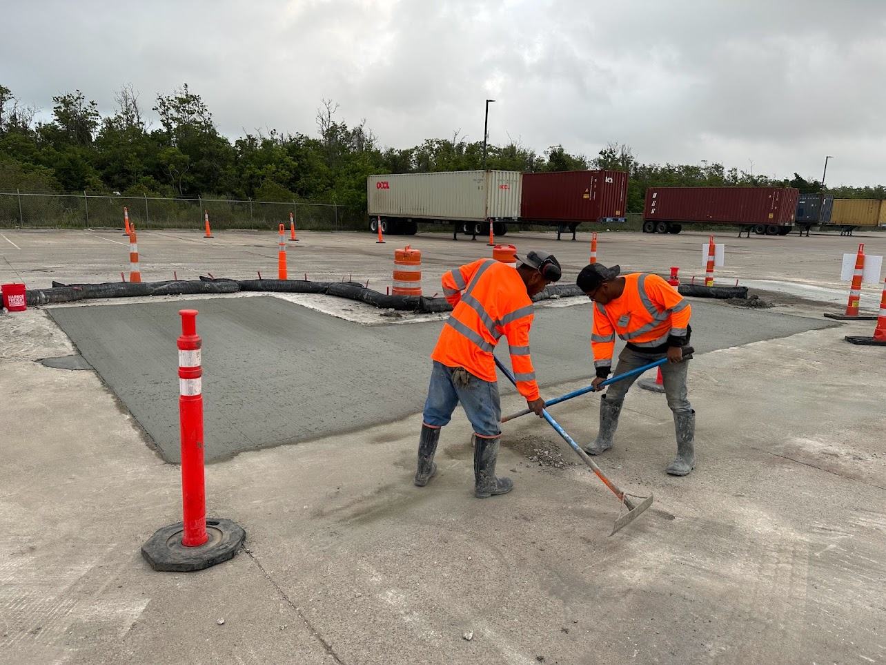Two construction workers in orange safety jackets smoothing wet concrete in a parking area surrounded by traffic cones and containers.