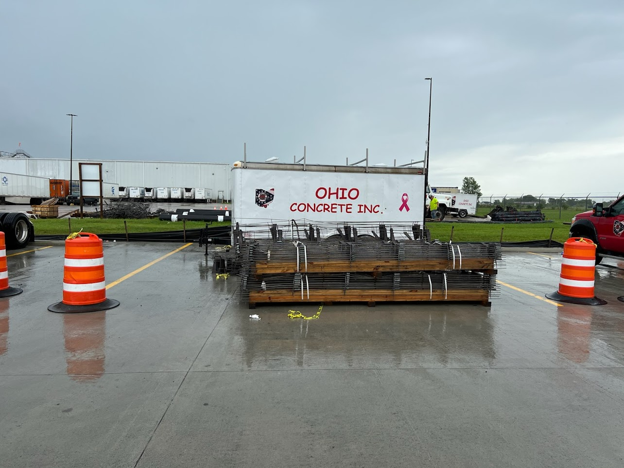 Concrete construction site on wet pavement with metal rebar stacks and a trailer displaying the Ohio Concrete Inc. logo and a pink ribbon symbol.