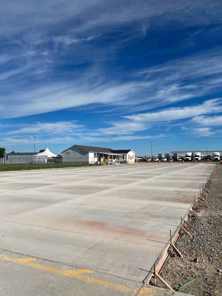 Wide concrete pavement area with construction stakes on the right edge, small buildings and trucks in the background under a partly cloudy blue sky.