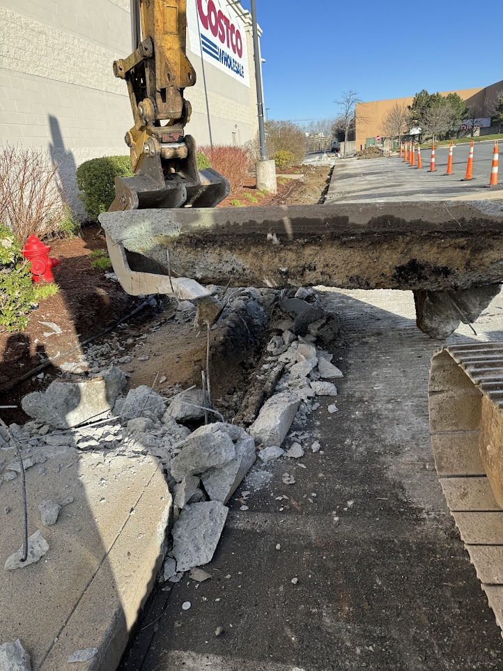 Excavator lifting broken concrete slab from a construction site outside a Costco Wholesale store with orange traffic cones along the road.
