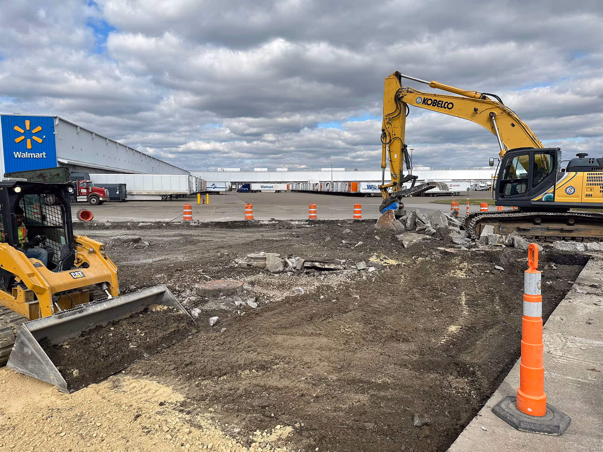 Construction site with excavator and bulldozer working on ground near Walmart distribution center under cloudy sky.