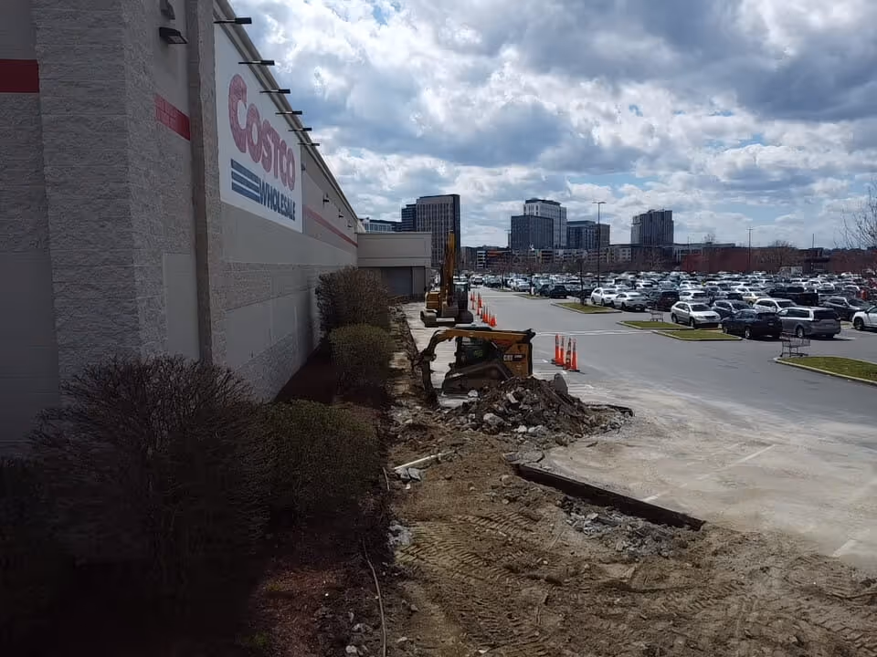 Construction equipment working on a sidewalk area outside a Costco Wholesale store with a large parking lot and city buildings in the background under a partly cloudy sky.