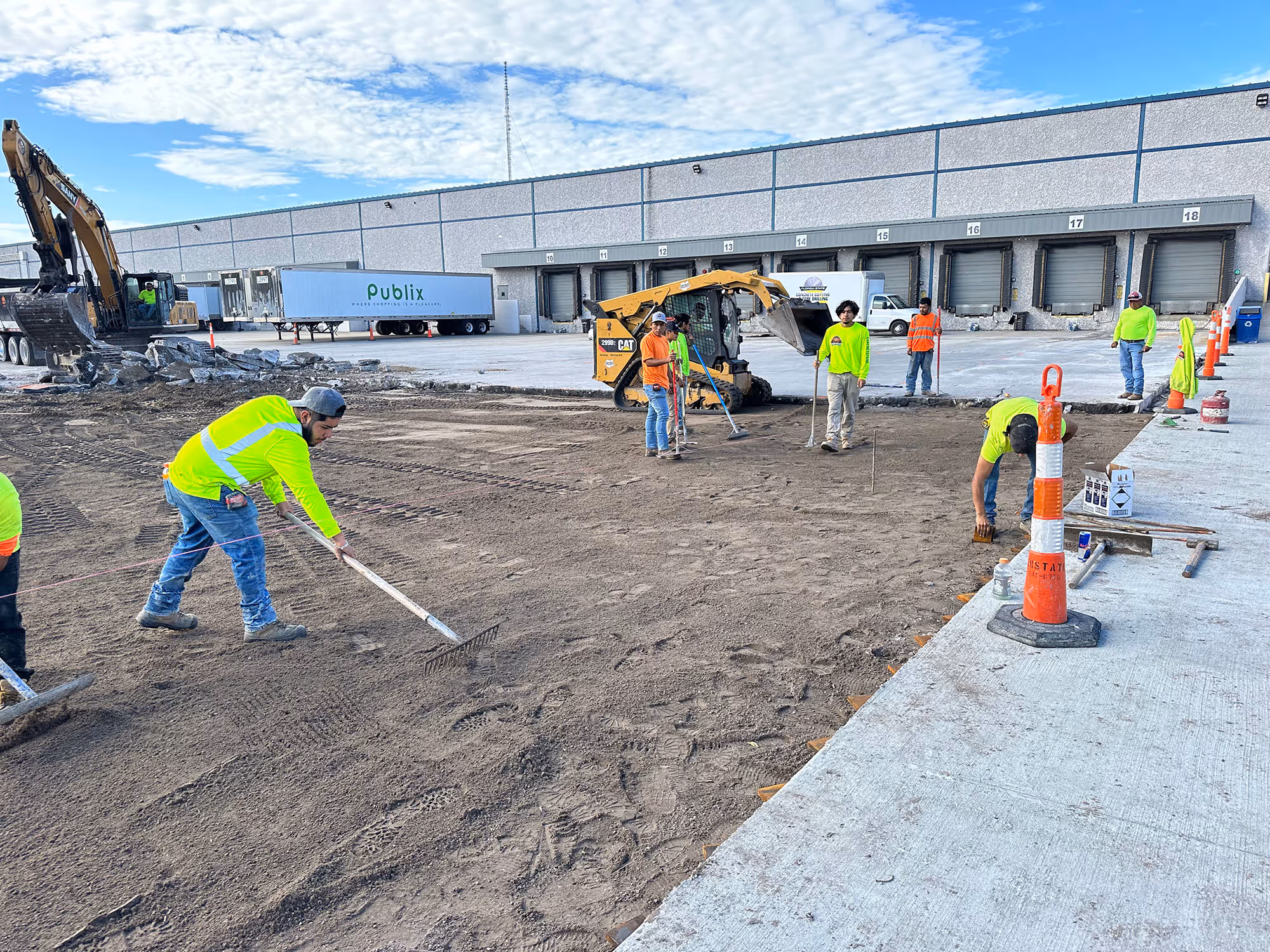 Construction workers in bright safety clothing leveling dirt and preparing ground at a commercial loading dock area with machinery and trucks in the background.