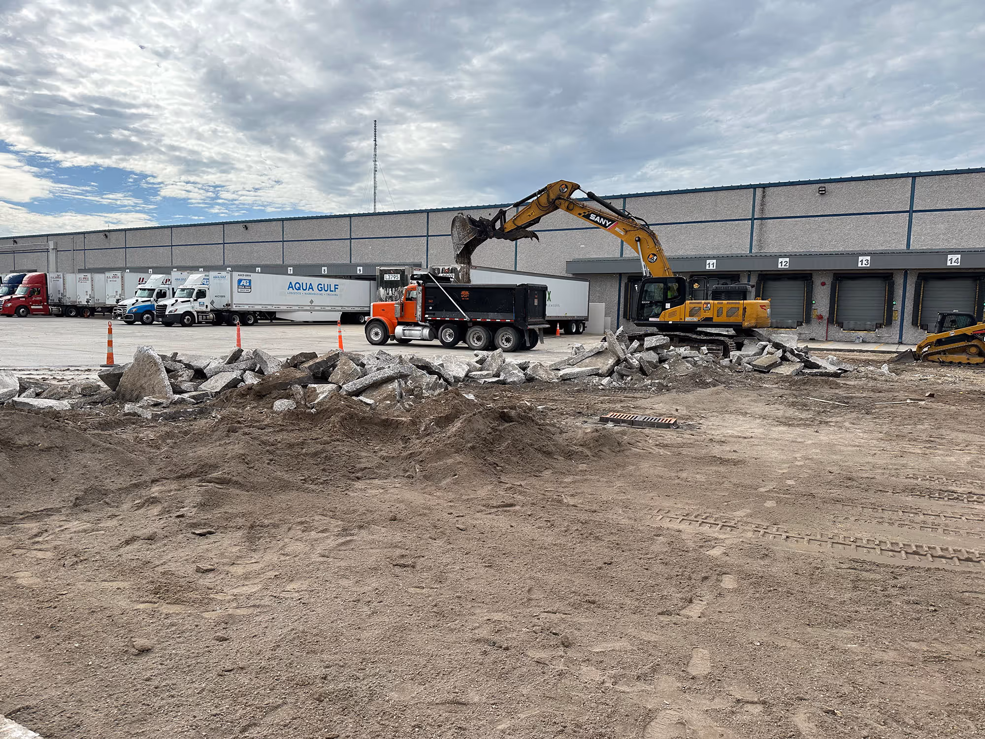 Construction site with excavator loading debris into a dump truck in front of a warehouse with parked trailers and delivery docks.