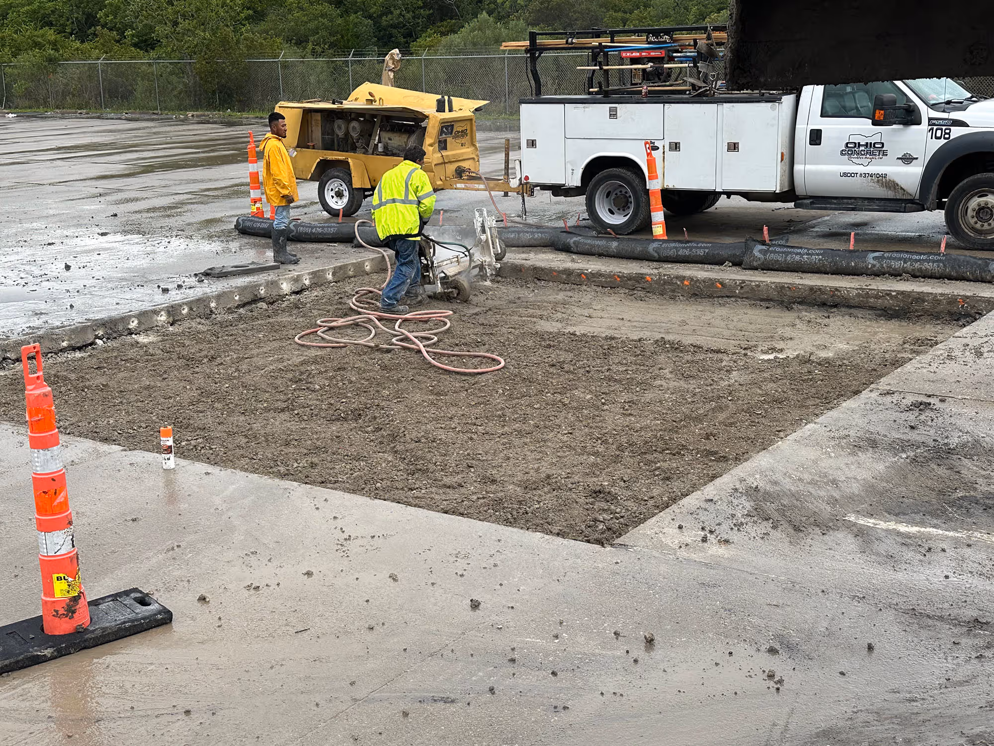 Two construction workers in rain jackets working on a concrete site with machinery and a white utility truck labeled Ohio Concrete.