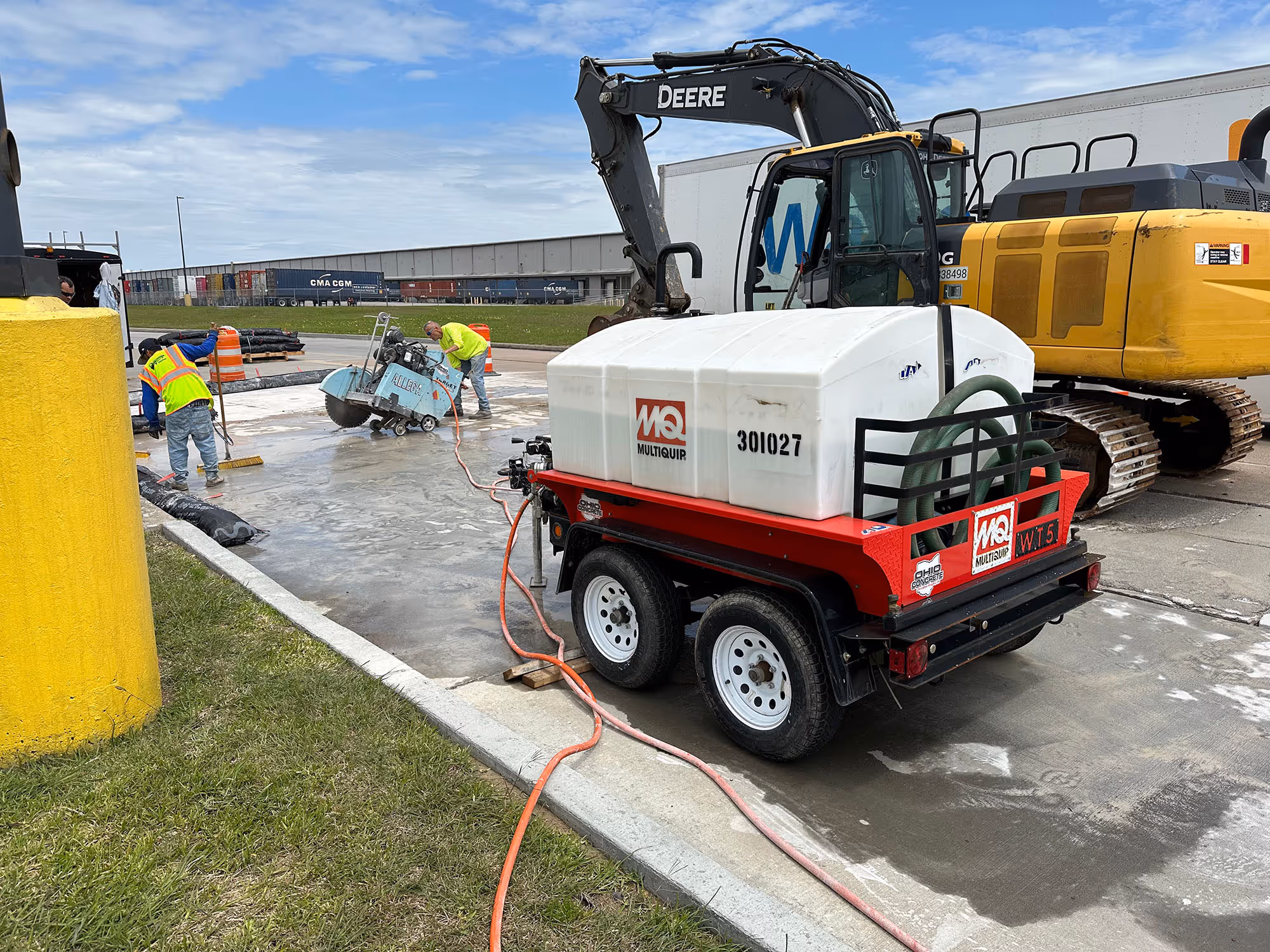 Construction workers using a concrete cutting saw on a wet pavement near a MultiQuip water tank trailer and a yellow John Deere excavator.