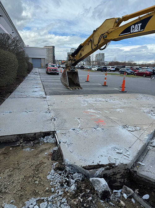 Construction site showing a CAT excavator digging up broken concrete sidewalk near a parking lot with orange traffic cones and parked cars in the background.