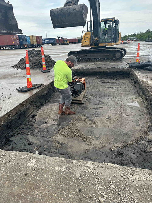 Construction worker operating compacting equipment inside a rectangular excavation site with an excavator in the background at a paved industrial area.