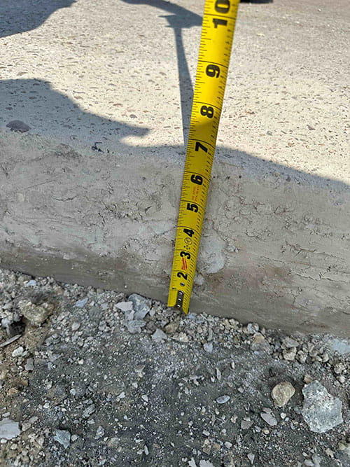Yellow measuring tape held vertically against a concrete edge showing a depth of about 5.5 inches.