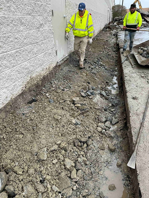 Two construction workers in yellow safety jackets walking beside a narrow trench filled with dirt and rocks next to a white brick building.