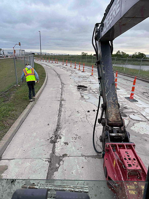 Construction worker in a high-visibility vest walking on cracked concrete road next to an excavator arm and orange traffic cones.