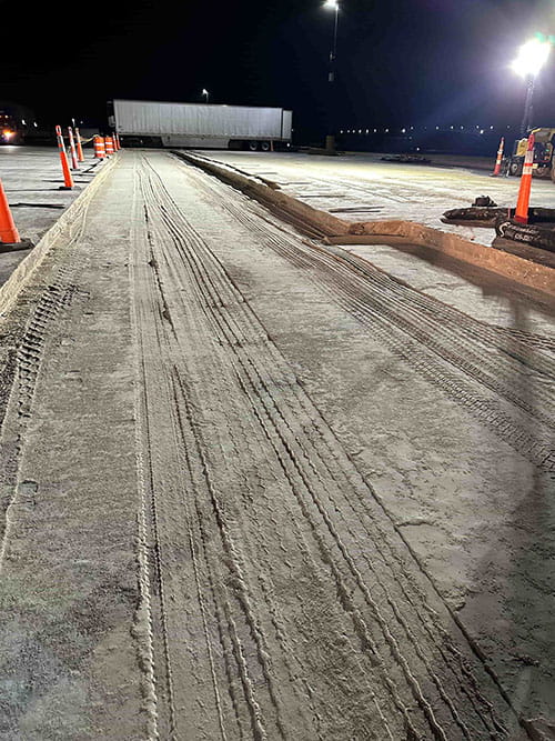 Nighttime construction site with freshly laid concrete marked by tire tracks and orange safety cones.