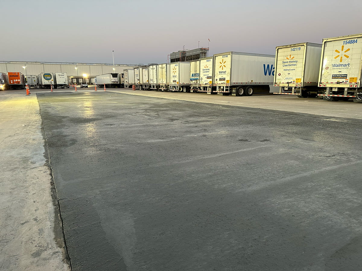 Parking area with freshly poured concrete and a row of Walmart delivery trucks docked at a warehouse during dusk.
