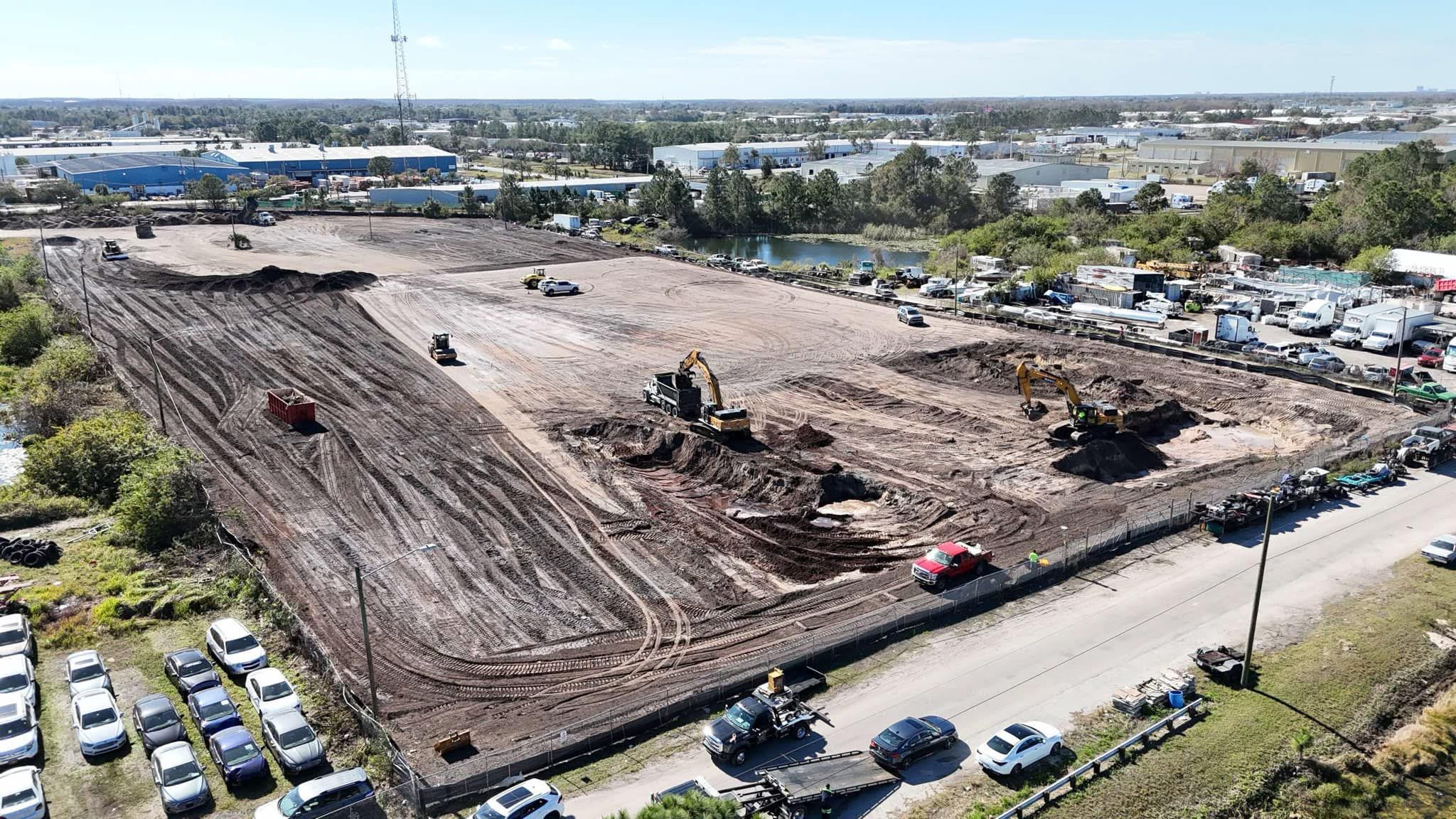 Aerial view of a construction site with excavators digging and trucks parked nearby, surrounded by industrial buildings and vehicles.