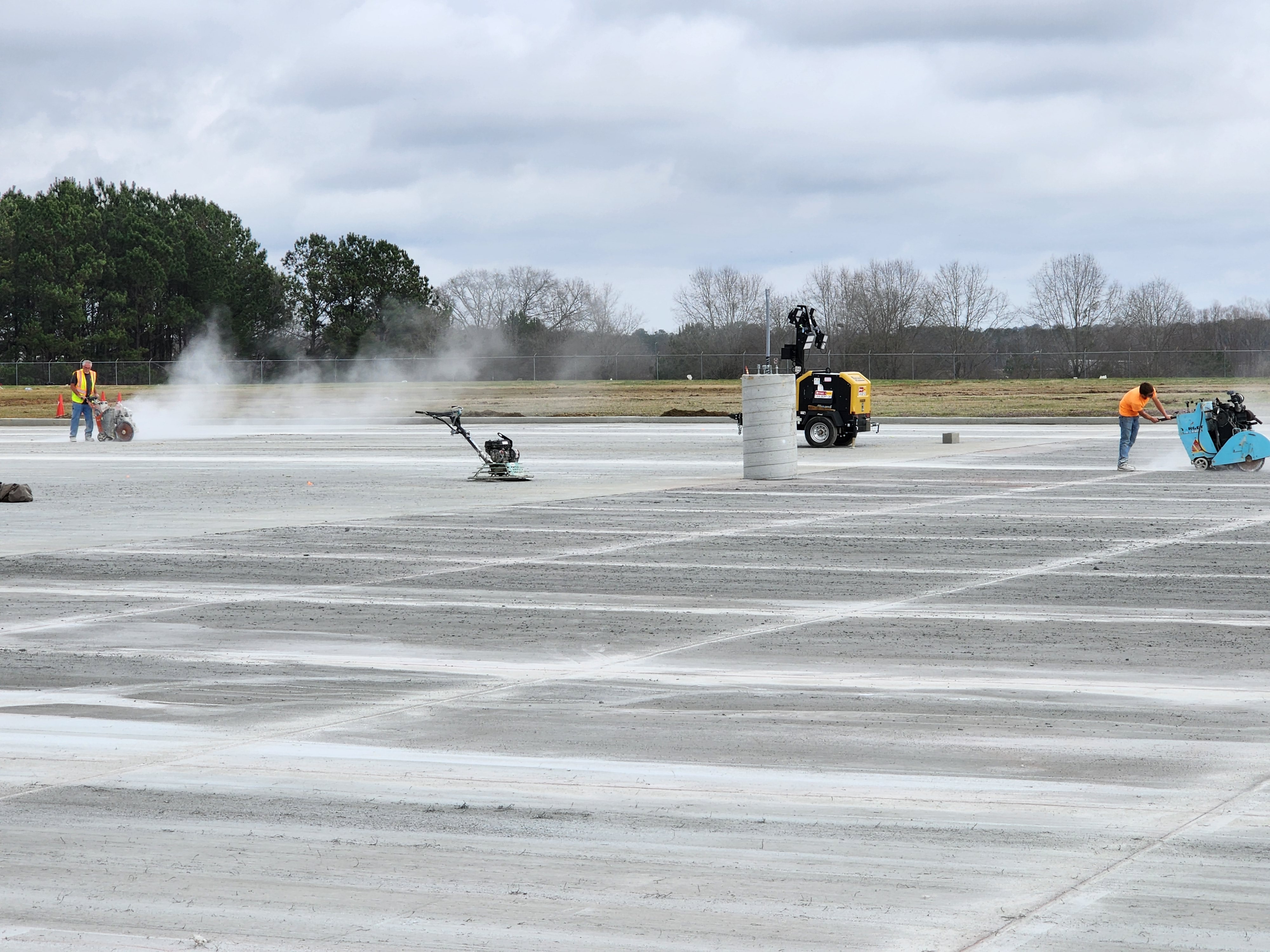 Workers using machinery to cut and smooth a large concrete surface on a cloudy day.