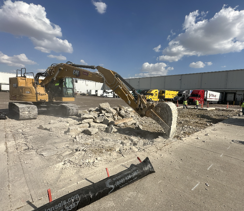 Yellow CAT excavator breaking concrete with rubble piles and trucks in an industrial yard under a blue sky with scattered clouds.