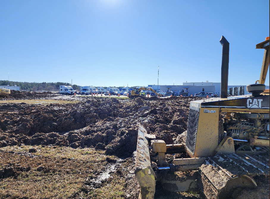 Construction site with a CAT bulldozer pushing dirt in the foreground and a row of trucks and buildings in the background under a clear blue sky.
