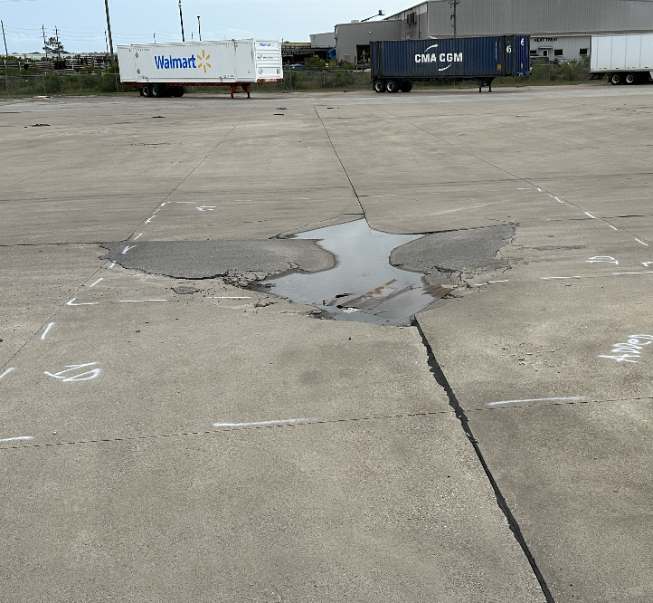 Large cracked pothole filled with water on a concrete surface in an industrial area with Walmart and CMA CGM shipping containers in the background.