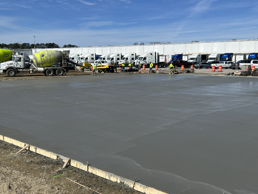 Construction workers in safety vests smoothing a large freshly poured concrete slab at an industrial site with cement mixer trucks and semi-trailers in the background.