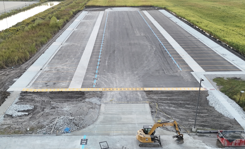 Aerial view of a large, newly paved parking lot under construction with marked spaces, adjacent grassy fields, and an excavator parked nearby.