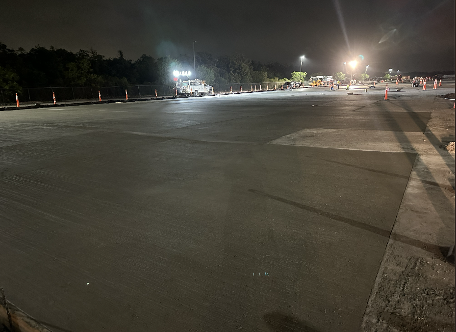 Night view of a large, freshly paved concrete surface with construction vehicles and orange safety cones along the edges.