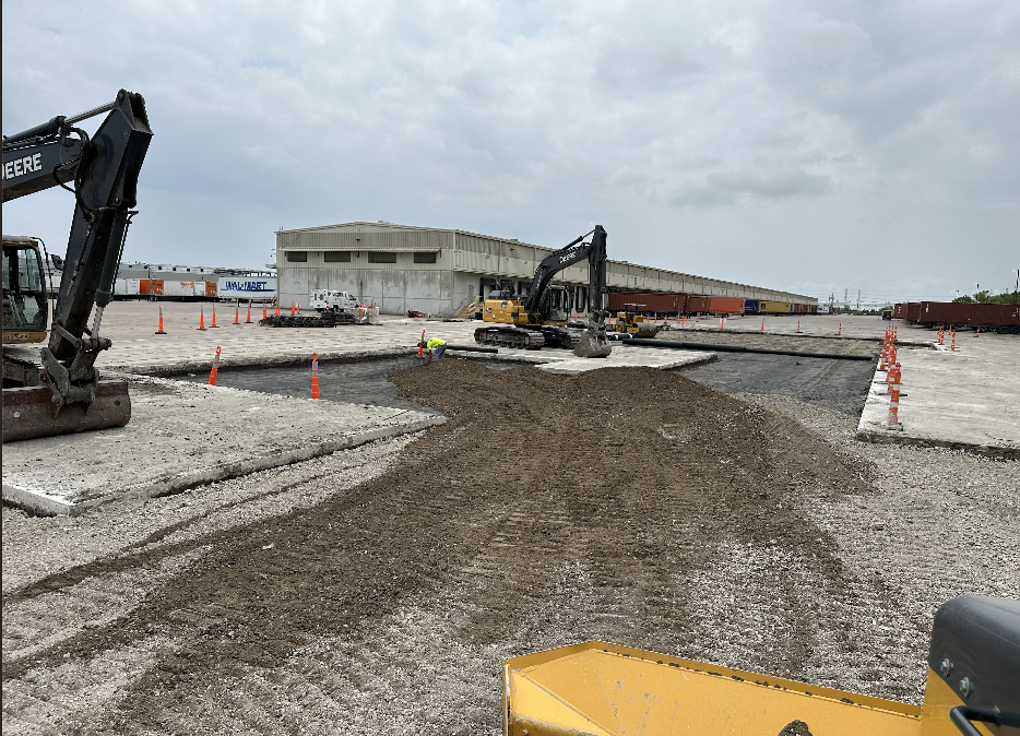 Construction site with excavators, dirt and gravel ground being leveled, and a large warehouse building in the background.