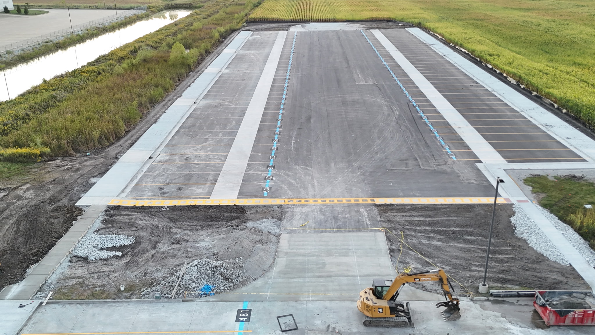 Large empty parking lot under construction with a yellow excavator and a red dumpster near the entrance, surrounded by grassy fields.