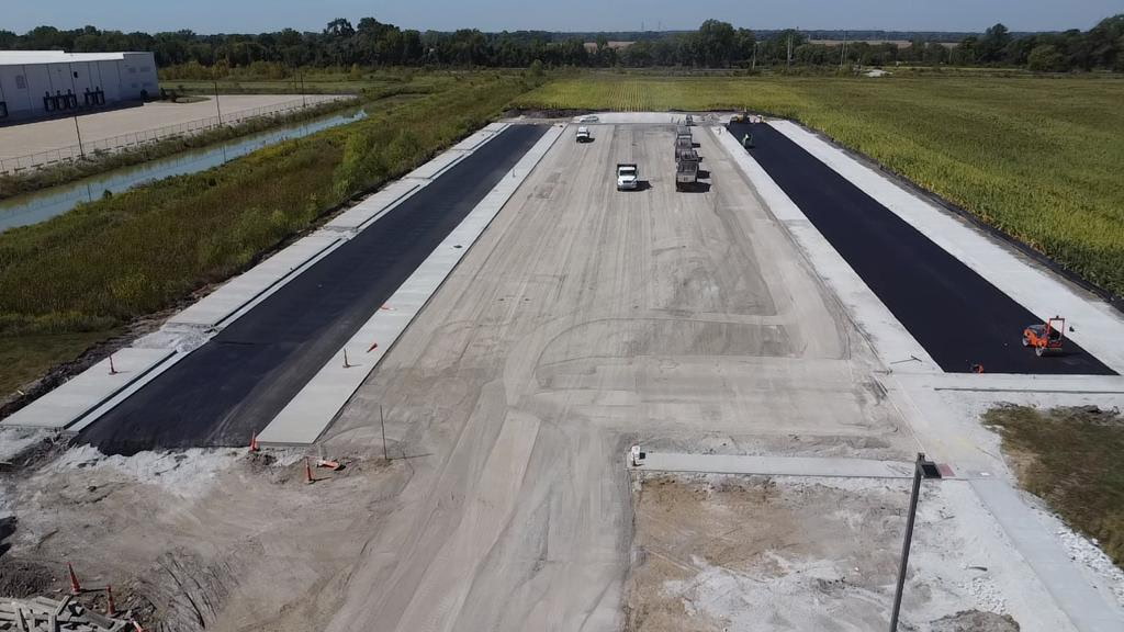Aerial view of a construction site with two newly paved black asphalt strips alongside concrete pathways, surrounded by green fields.